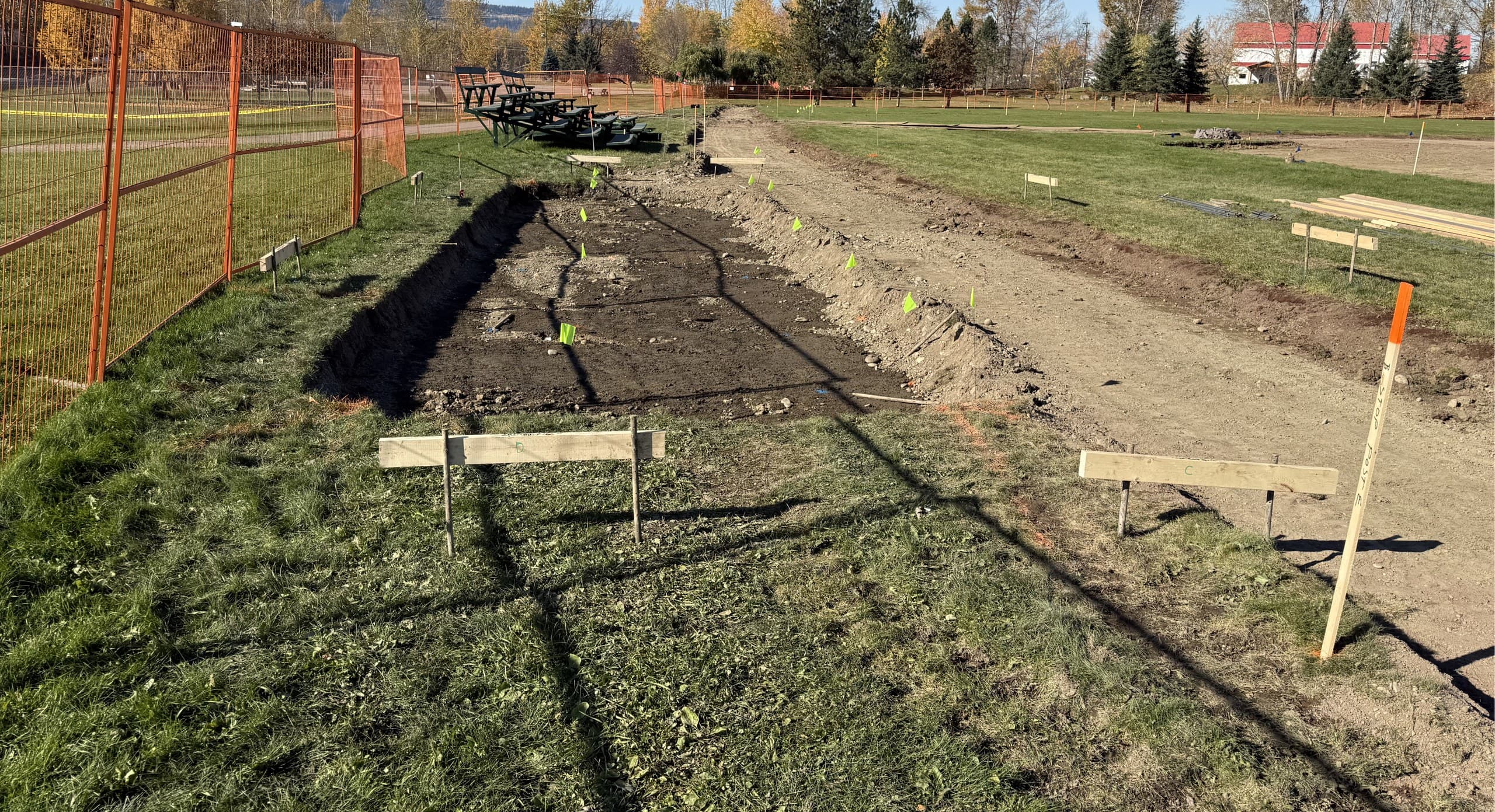 Servicing trench laid out with stakes and offset boards beside orange construction fencing